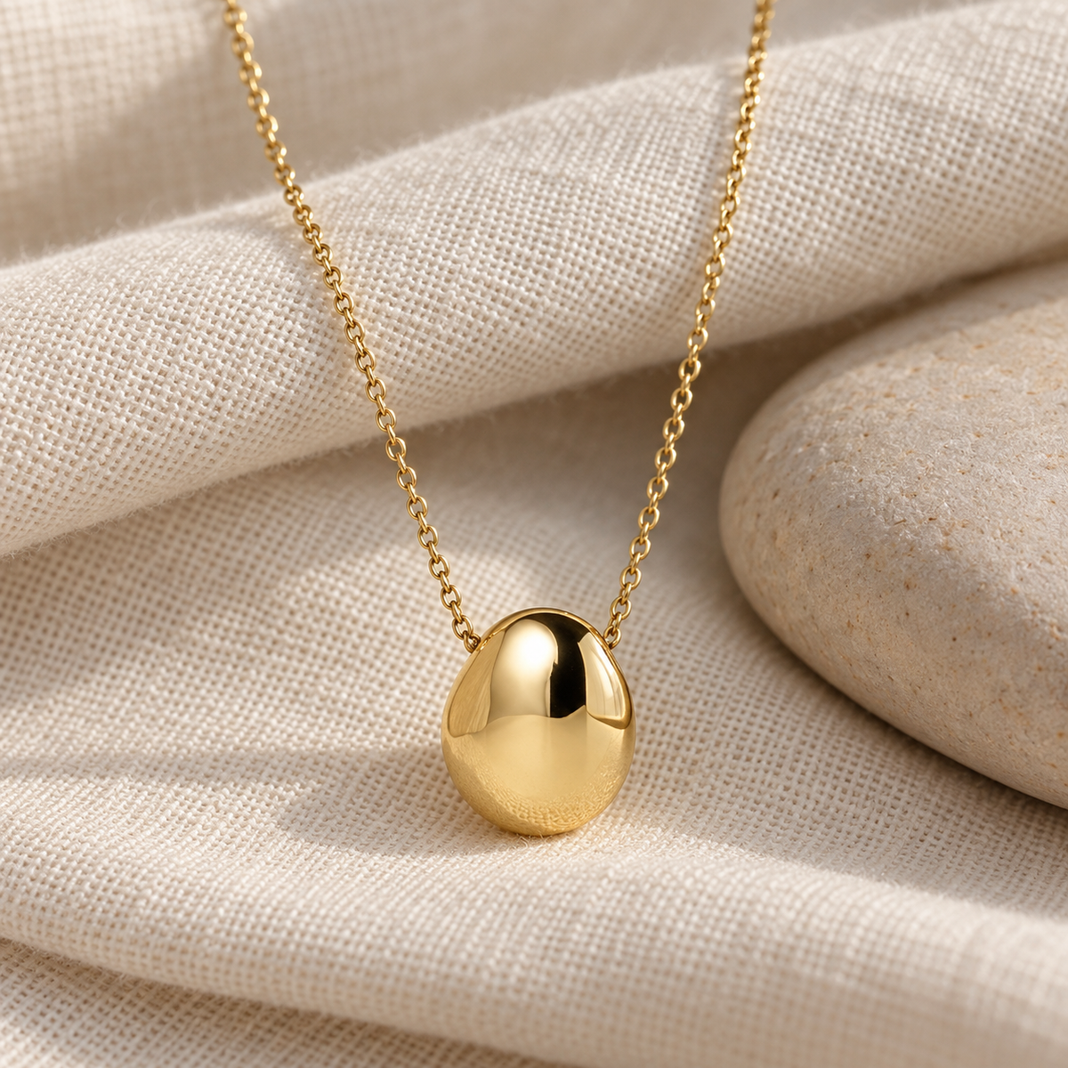 Gold Pebble Pendant Necklace on natural linen beside a pale stone